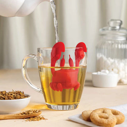 Red lobster-shaped tea infuser in a glass of tea with water being poured, on a table with tea leaves and cookies.