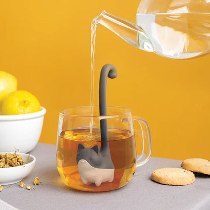 Tea being poured into a glass mug with a cat-shaped tea infuser against a yellow background.