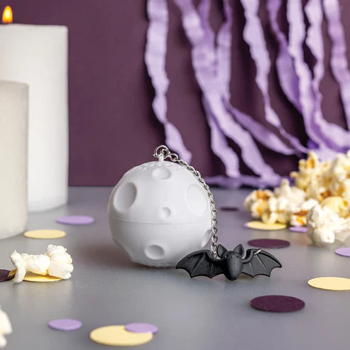 White spherical Halloween-themed tea infuser with a bat charm on a table with popcorn and candles.